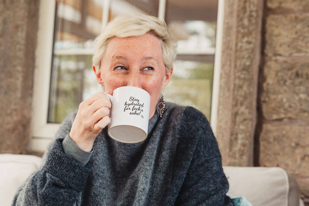 Nic Redman, voice coach in UK, happily drinking in a mug with words printed on it saying stay hydrated for feck's sake.