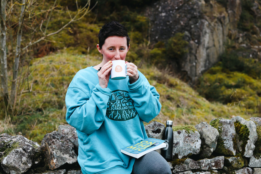 Nic sitting on a wall in a blue jumper enjoying a cup of team, reading her book On The Mic.